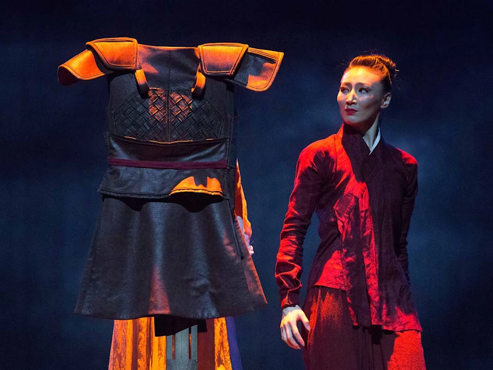 A woman in a red outfit stands on stage next to a traditional armor costume displayed on a stand, with dramatic lighting and a dark background.