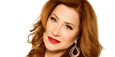 A woman with long, wavy red hair and green eyes is looking at the camera, wearing a black top and gold hoop earrings.