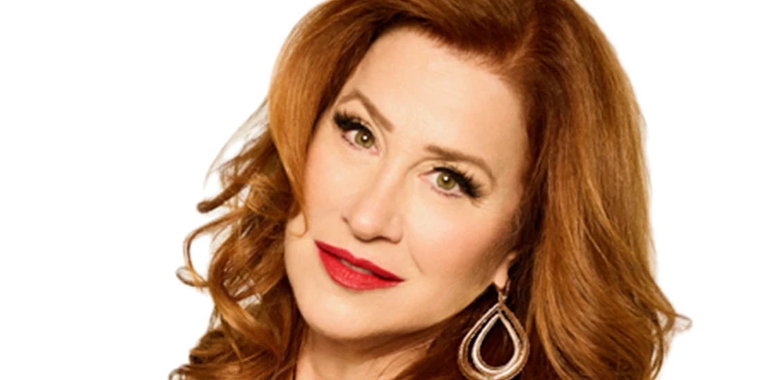 A woman with long, wavy red hair and green eyes is looking at the camera, wearing a black top and gold hoop earrings.