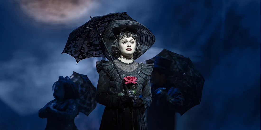 A woman in dark Victorian-style clothing holds a red rose and a black umbrella on stage, with two people holding umbrellas in the background under blue lighting.