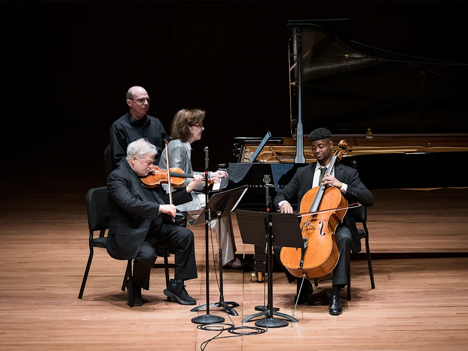 Four musicians perform on stage: a violinist, pianist, cellist, and page turner. A grand piano is in the background, and music stands are in front of the seated musicians.
