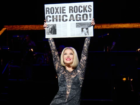 A performer on stage (Ariana Madix), dressed in a black lace outfit, holds up a newspaper with the headline "ROXIE ROCKS CHICAGO!.