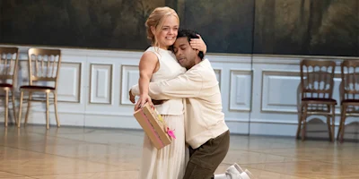 A woman in a white dress smiles while being hugged by a man kneeling on the floor; she holds a gift box and they stand in a room with wooden chairs and paneled walls.
