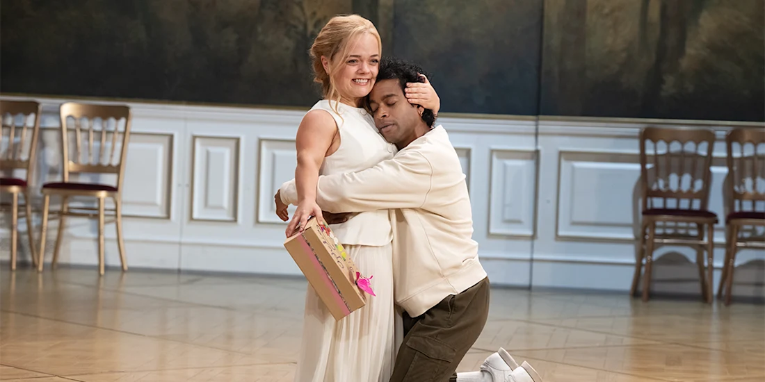 A woman in a white dress smiles while being hugged by a man kneeling on the floor; she holds a gift box and they stand in a room with wooden chairs and paneled walls.