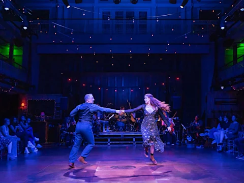 A man and woman dance together on stage holding hands, with a live band playing behind them and an audience watching from either side under warm string lights.