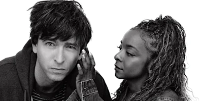 A man looks directly at the camera while a woman stands beside him, appearing to whisper in his ear against a plain white background.