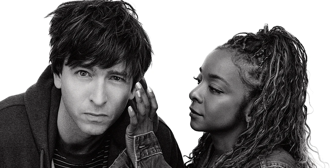 A man looks directly at the camera while a woman stands beside him, appearing to whisper in his ear against a plain white background.