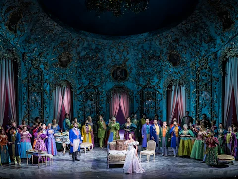 A large group of performers in colorful costumes stand on an ornate stage with elaborate blue and gold décor, gathered around a woman in a white gown at the center.