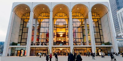 People gather outside the grand glass and stone entrance of Lincoln Center’s Metropolitan Opera House in New York City during the evening.