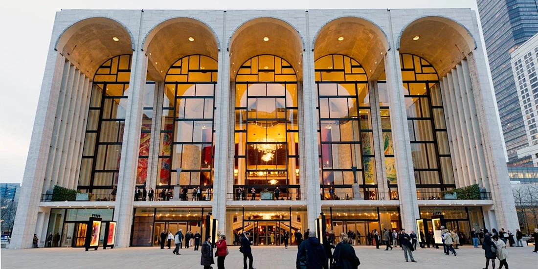 People gather outside the grand glass and stone entrance of Lincoln Center’s Metropolitan Opera House in New York City during the evening.