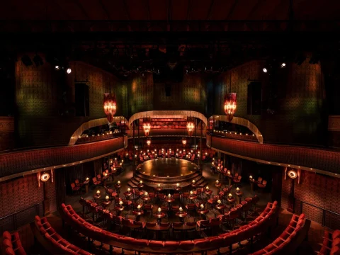 A dimly lit, ornate theater with red velvet seats and a circular stage at the center. The ambiance is warm, with decorative chandeliers and detailed architectural design.