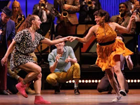 Two women dance joyfully on stage with a jazz band playing in the background. A group of musicians watches, and a kneeling man in the center claps enthusiastically.