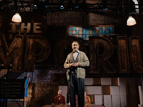 An older man stands and speaks on stage in front of large letters spelling “THE EMPORIUM,” with two people seated behind him and theatrical lighting overhead.