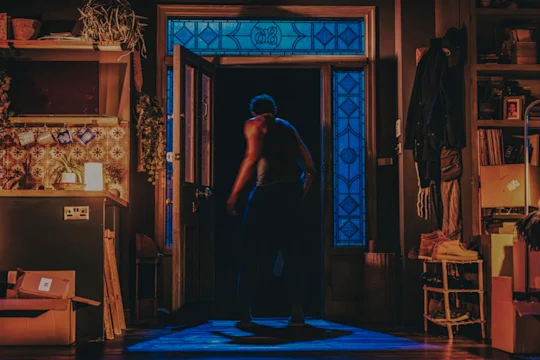 A man stands silhouetted in an open doorway as blue light pours across a cluttered, boxed-up living room.