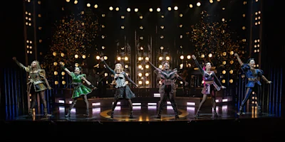 Six women in colorful, Tudor-inspired costumes perform on stage under bright lights, with confetti falling above them.