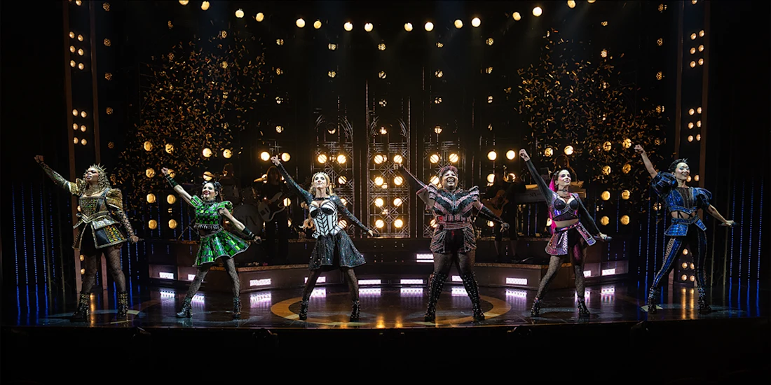 Six women in colorful, Tudor-inspired costumes perform on stage under bright lights, with confetti falling above them.