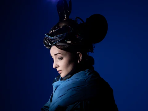 A person wearing a blue coat and an elaborate headpiece with wires and objects, set against a dark blue background.