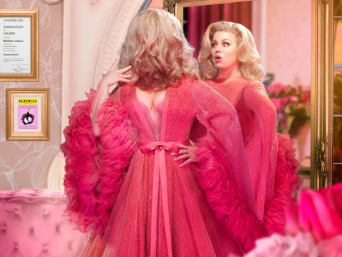 A person with styled blonde hair looks at themselves in a mirror, wearing a pink gown with feathered sleeves in a well-decorated room.