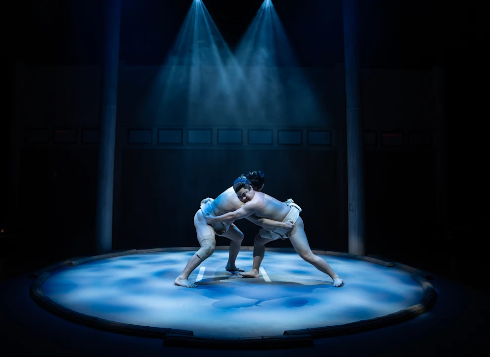 Two sumo wrestlers engaged in a match under focused stage lighting, surrounded by a circular ring and atmospheric smoke.