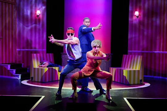 Three actors on stage strike dramatic poses, one in an orange suit crouching, one in a tie and red mask, and one in a blue suit standing upright; set has striped walls and chairs.