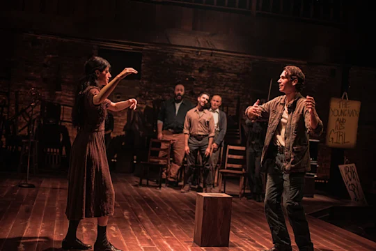 A woman and a man face each other on a wooden stage, with three other men standing in the background.