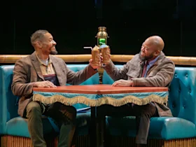 Two people sitting on a blue booth bench raise drinks with straws in a celebratory toast, smiling at each other over a table with a fringed edge.