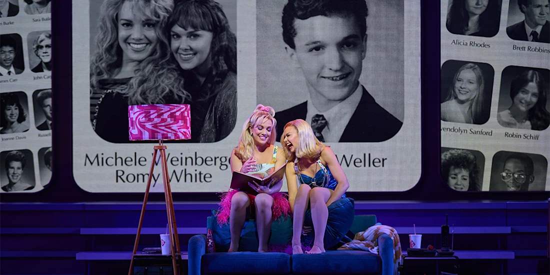 Two women sit on a couch looking at a yearbook, while large black-and-white high school portraits are projected behind them on stage.