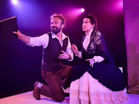 A man kneels and extends a book while a seated woman in period costume looks on under purple stage lights.