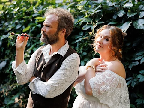 A man holding a long cigarette holder stands next to a woman in a white lace dress, both posing outdoors in front of dense green foliage.
