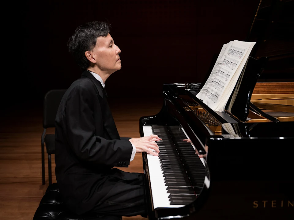A person in formal attire plays a grand piano on stage, reading sheet music.