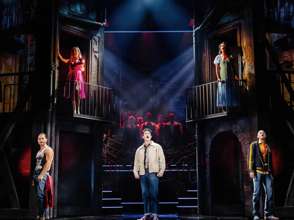 Five actors perform on stage, three on elevated balconies and two on the ground, amidst a dramatic urban backdrop with spotlights highlighting each performer.