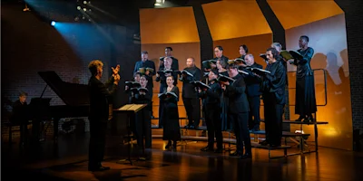 A conductor leads a choir of singers holding music folders on a stage with a grand piano.