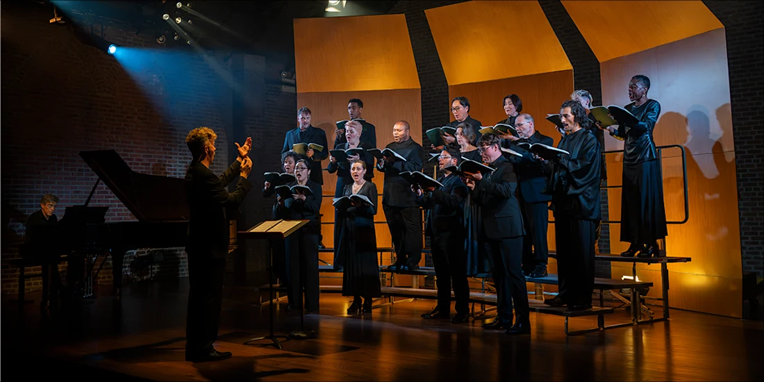 A conductor leads a choir of singers holding music folders on a stage with a grand piano.