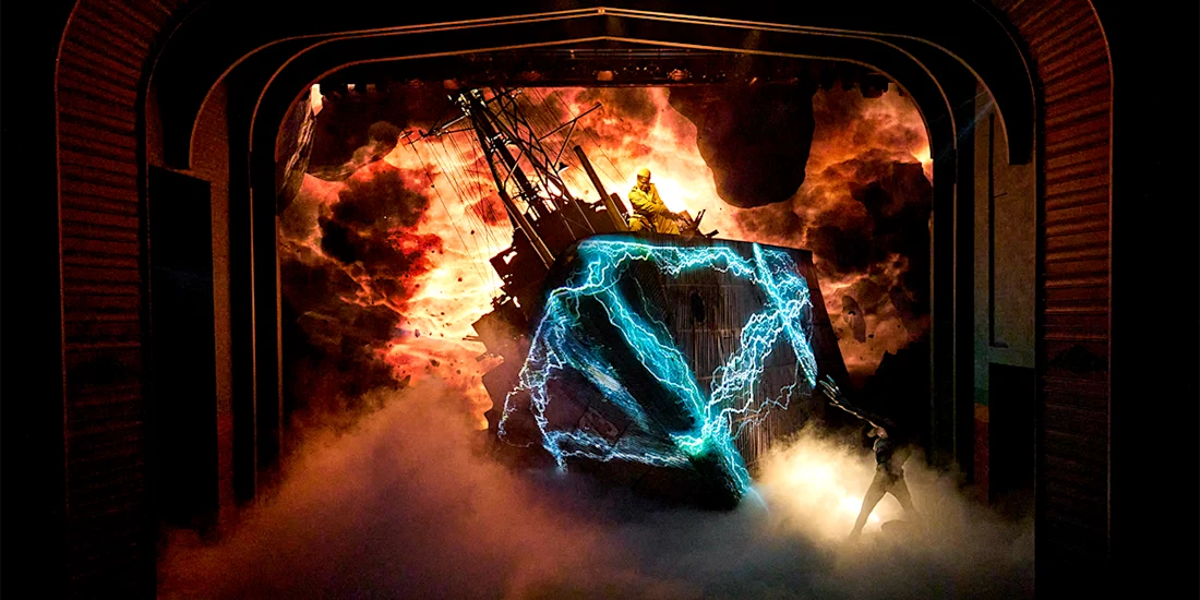 A dramatic stage scene with smoke, lightning effects, a shipwreck set, and a person standing atop the wreck amid an intense, fiery background.