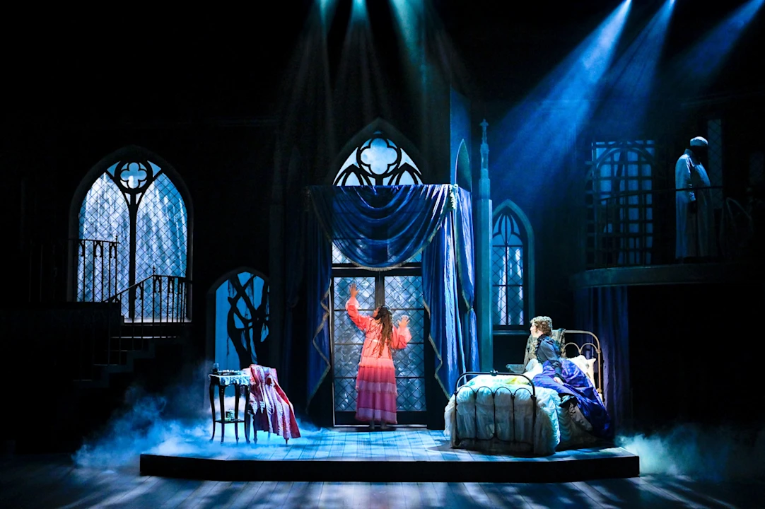 A theater stage showing a woman in a pink dress standing at a window and another person sitting on a bed, with a silhouette in an upper balcony.