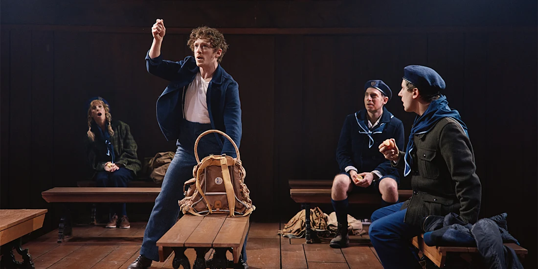 Four people in scout uniforms sit on wooden benches indoors; one person stands holding up an apple, while others watch or eat apples, with backpacks on the floor.