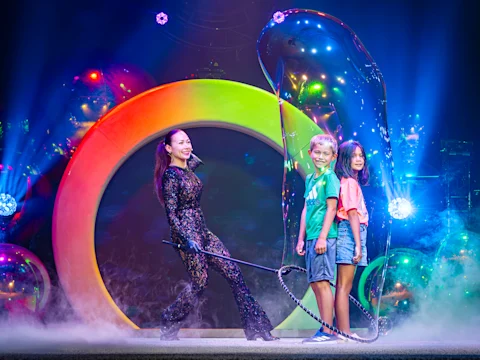 A performer in a black outfit creates a large bubble around two smiling children on stage, with colorful lights and smoke effects in the background.