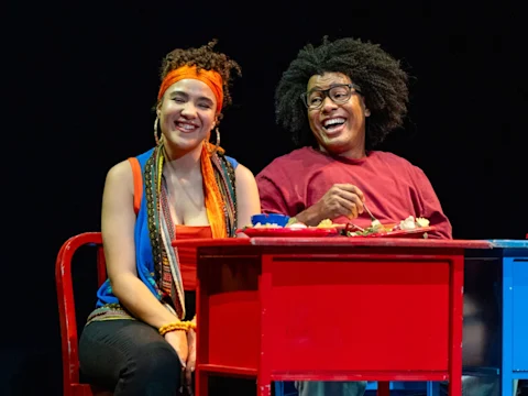 Two people sit at a red table, smiling and laughing, with food and drinks in front of them against a dark background.
