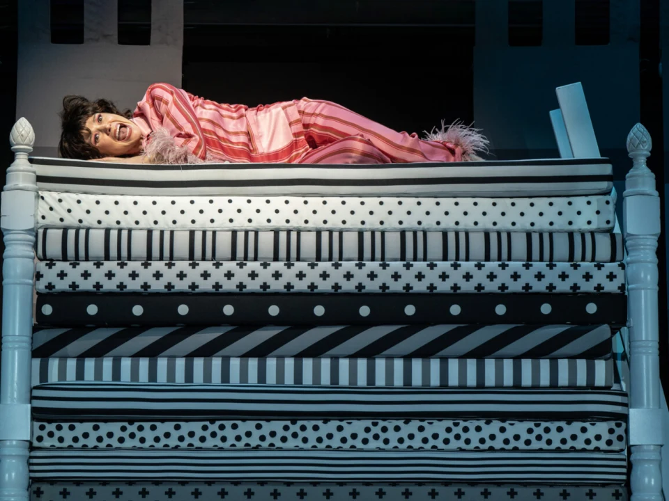 A person lies on a tall stack of mattresses, wearing striped pajamas, in a theatrical setting.