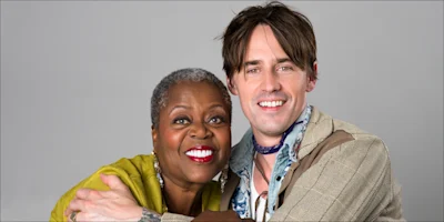 Two people, one older woman with short gray hair and one younger man with brown hair, smiling and hugging in front of a plain gray background.
