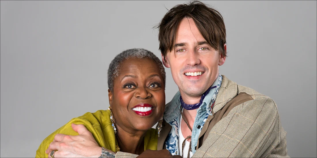 Two people, one older woman with short gray hair and one younger man with brown hair, smiling and hugging in front of a plain gray background.