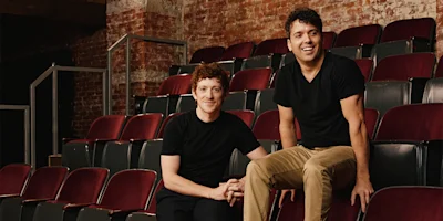 Six people pose in an empty theater with red seats and exposed brick walls, five standing or sitting behind a seated woman in black overalls at the front.