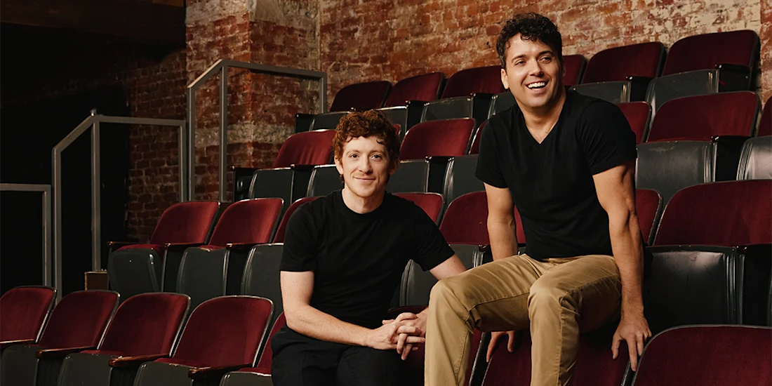 Six people pose in an empty theater with red seats and exposed brick walls, five standing or sitting behind a seated woman in black overalls at the front.