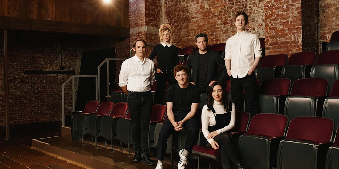 Six people pose in an empty theater with red seats and exposed brick walls, five standing or sitting behind a seated woman in black overalls at the front.