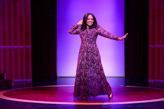 A woman in a long purple patterned dress poses and smiles on a circular stage with a purple-lit backdrop.