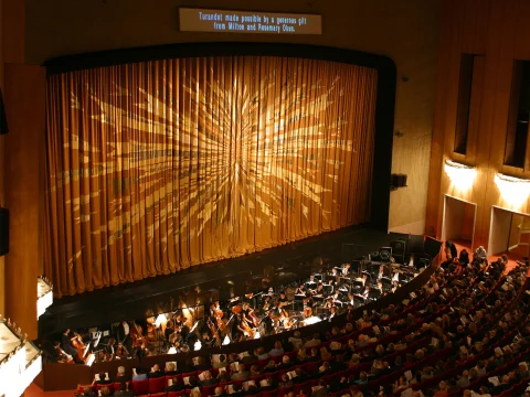 Production photo of Renee Fleming In Recital in Los Angeles showing, Orchestra performing on stage in a theater with a large, radiant sunburst design curtain and an audience watching in red seats.