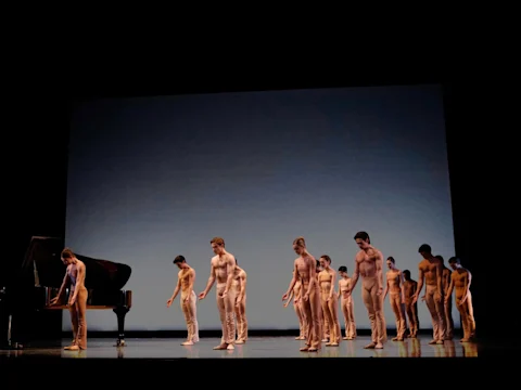 A group of male dancers in skin-toned costumes stands in a line on stage beside a grand piano, performing under a plain blue-gray backdrop.