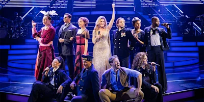 A group of performers in costumes sings on a brightly lit stage, with one woman in a gold dress standing at the center raising her arm.