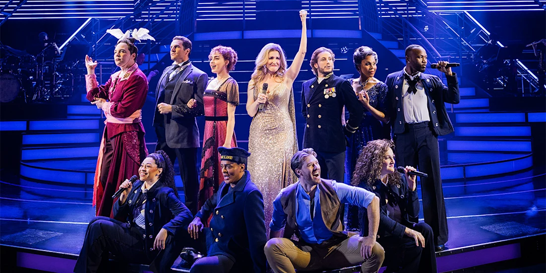 A group of performers in costumes sings on a brightly lit stage, with one woman in a gold dress standing at the center raising her arm.