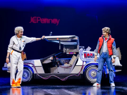 Don Stephenson as Doc Brown, Caden Brauch as Marty McFly stand beside a DeLorean car on stage, one dressed in a white jumpsuit and the other in a red puffy vest. A JCPenney sign is visible in the background.
